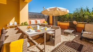 Une terrasse extérieure aménagée avec des meubles en osier, une table dressée avec des assiettes, des verres et une carafe, et un parasol pour faire de l’ombre.