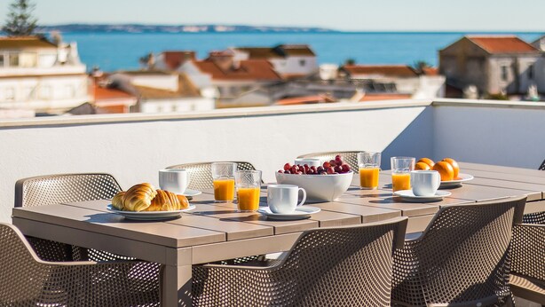 Tavola per la colazione con croissant, frutta e succo, con vista su una cittadina costiera.