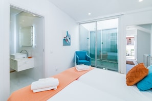 Ein modernes Schlafzimmer mit einem Bett, einem blauen Sessel, einer Glastür, die auf einen Balkon führt, und einem Badezimmer mit Spiegel und Waschbecken.