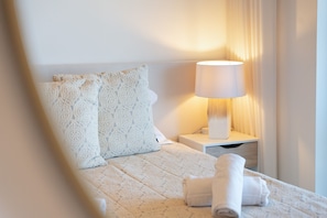 Een keurig opgemaakt bed met een witte deken en twee gestreepte kussens, een bedlampje en een opgerolde handdoek op een houten nachtkastje.