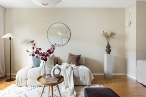 Ein modernes Wohnzimmer mit einer Couch, einem runden Spiegel, einer Blumenvase, einer Stehlampe und einer hohen Vase auf einem Sockel.