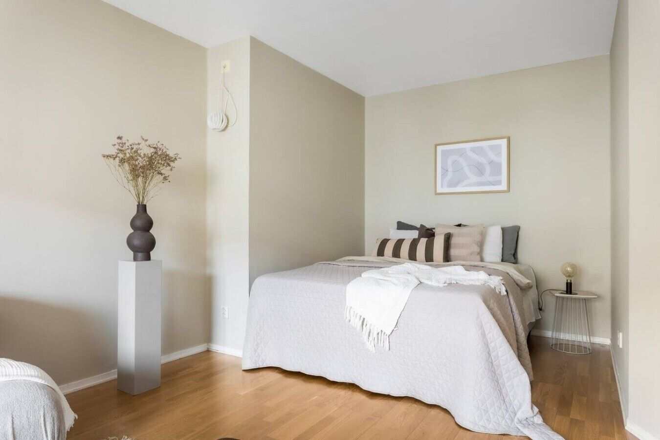 A bedroom with a bed, bedside table, lamp, and a vase with dried flowers.