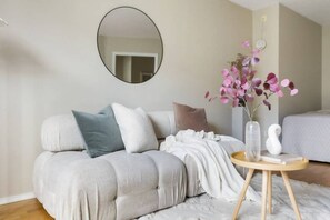 Un dormitorio moderno con un sofá capitonado, un espejo redondo, un jarrón con flores y una mesita de noche.