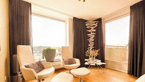 Une chambre moderne avec une grande fenêtre, une chaise blanche ornée d’un coussin à motifs léopard, un petit pouf blanc et un élément décoratif mural.