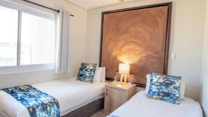 Habitación de hotel con dos camas, cabecera de madera, mesita de noche con lámpara y ventana con cortinas.