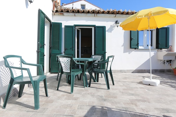 Une terrasse avec une table et des chaises, un parasol jaune et un bâtiment blanc aux volets verts.