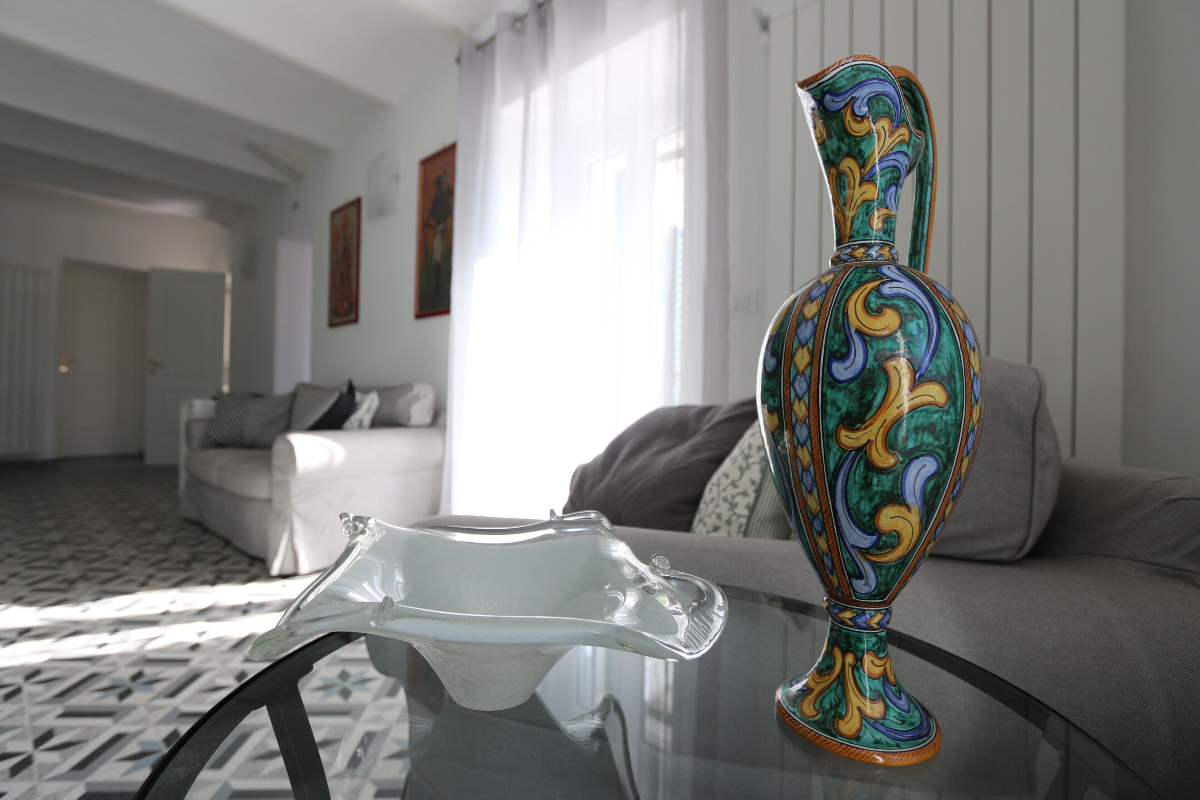 A room with a glass table, a colorful vase, a white sofa, and a patterned floor.