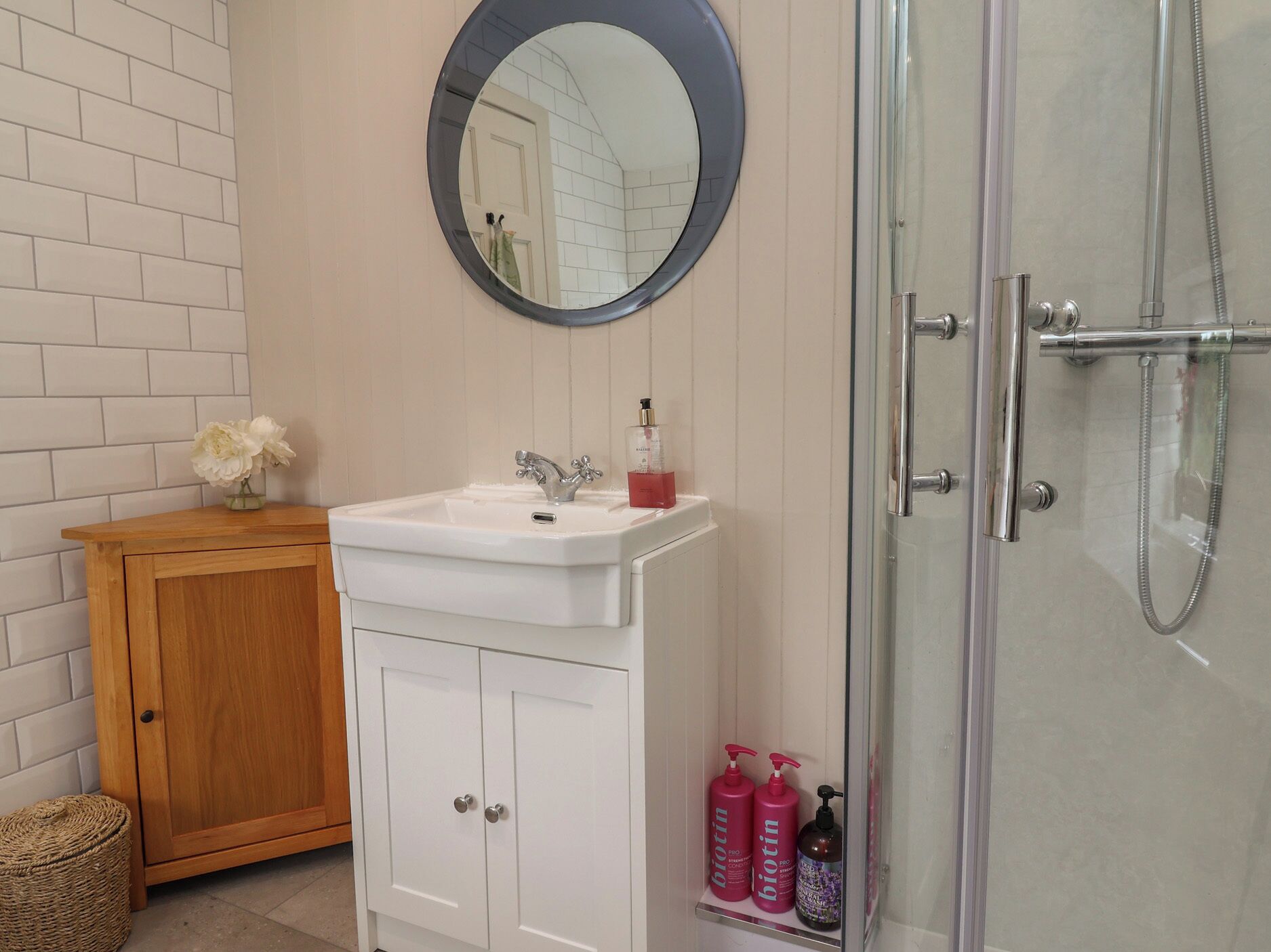 A bathroom with a white sink, a wooden cabinet, a round mirror, and a glass shower enclosure.