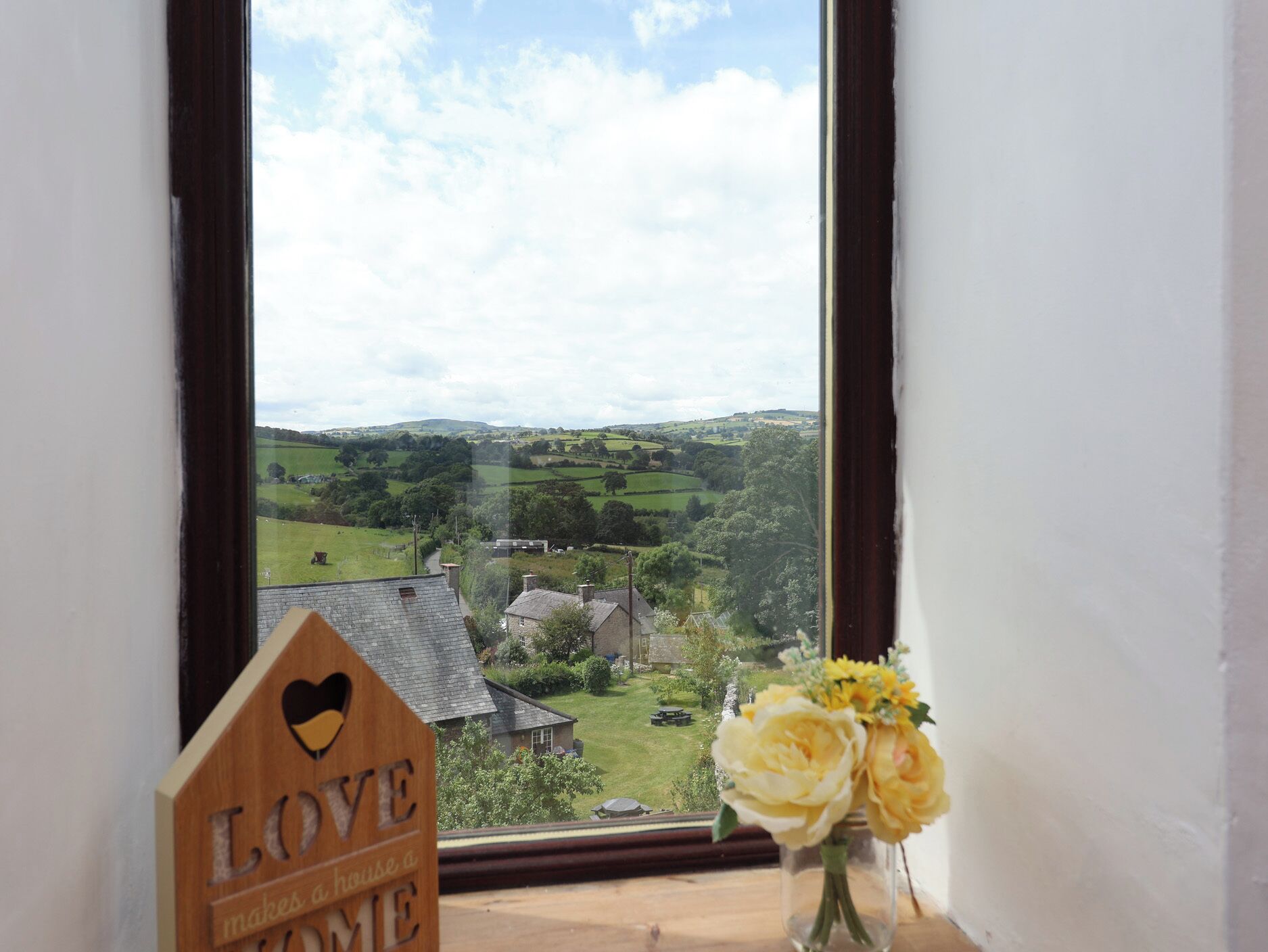 A view through a window with a wooden sign saying 'LOVE makes a home' and a vase with yellow flowers.