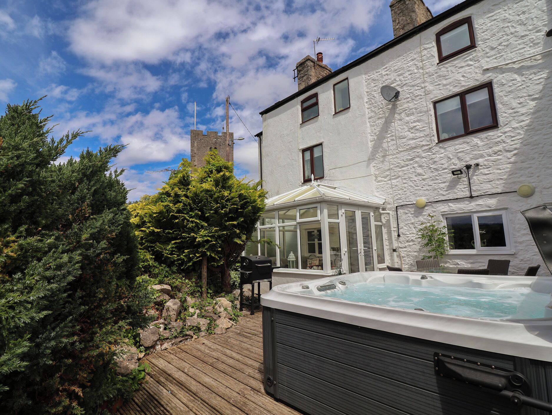 A hot tub in a backyard with a wooden deck, a white building, and a stone tower in the background.