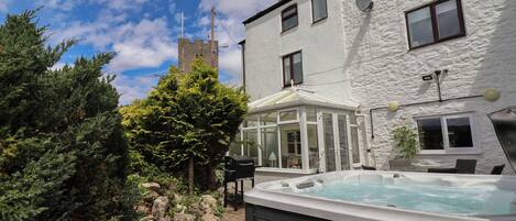 A hot tub in a backyard with a wooden deck, a white building, and a stone tower in the background.