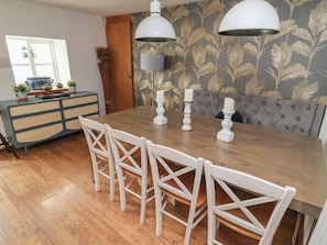 Un espace repas comprenant une table en bois, des chaises blanches, deux lampes suspendues et un banc recouvert d’un tissu gris.