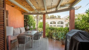 En udendørs terrasse med et bord og stole, grill med tag og udsigt til en bygning i baggrunden.