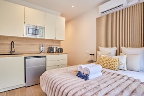 Ein modernes Hotelzimmer mit Küchenzeile, einem Bett mit flauschiger Decke und einem Nachttisch mit Büchern und Lampe.