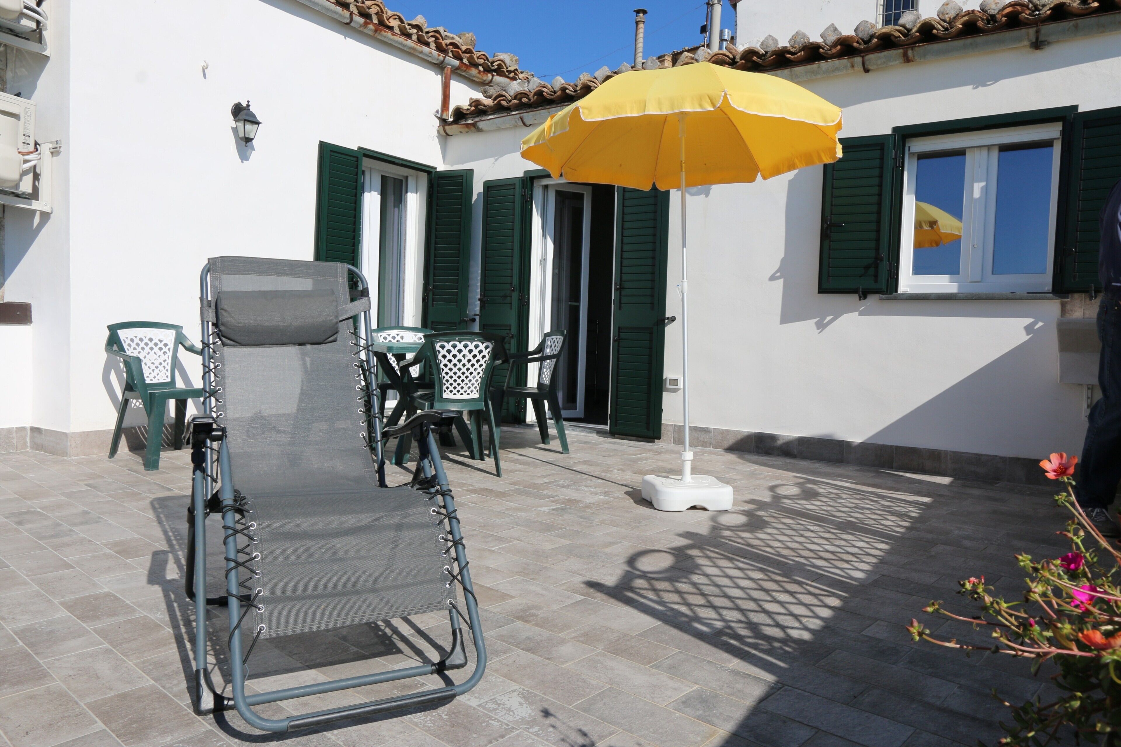 Un área de estar exterior con acceso para sillas de ruedas, una mesa, sillas, un paraguas amarillo y una planta en maceta.