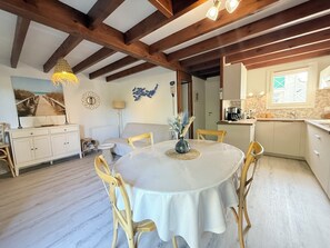Ein Zimmer mit einem runden Tisch, Holzsesseln, einem Bett, einem kleinen Beistelltisch, einer Lampe, einem Gemälde und einer Küchenzeile mit einem Fenster.