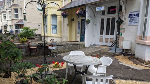Ein Hotel mit einer Terrasse, ausgestattet mit einem runden Tisch und weißen Stühlen, einem kleinen Garten mit Blumen und einem Schild, das den Namen und die angebotenen Leistungen des Hotels anzeigt.