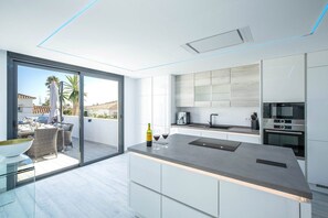 A modern kitchen with a large island, a built-in oven, and a glass door leading to a balcony with outdoor furniture. - Villa Las Farolas - El Faro Luxury Villa (Mijas Costa)