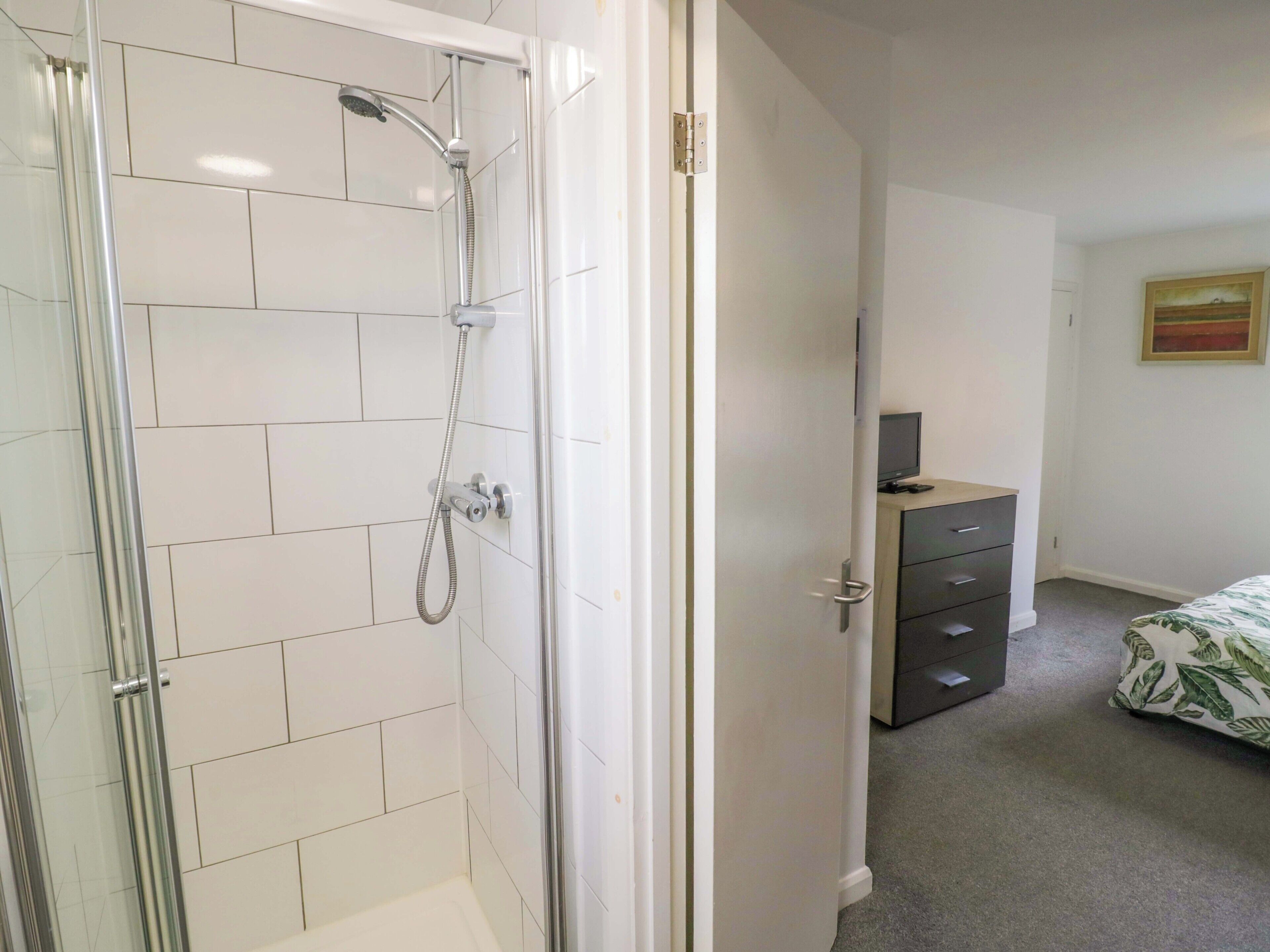 Une salle de bain avec une douche à parois de verre, des murs recouverts de carreaux blancs, une commode en bois et un lit avec une couverture à motifs de feuilles.