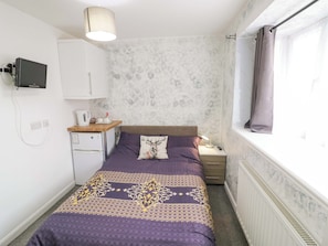 Ein kleines, ordentlich eingerichtetes Schlafzimmer mit Bett, Nachttisch, Lampe und einem Fenster mit Vorhängen.