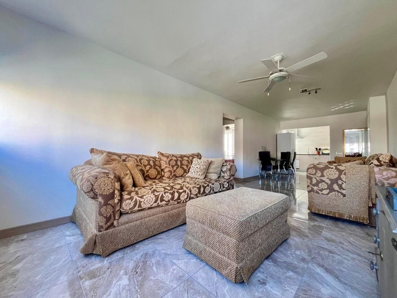A spacious living area with a sofa set, ottoman, ceiling fan, and a dining area in the background.