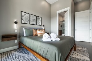 A bedroom with a bed, wall-mounted artwork, a bedside table, and a door. - Elevated Escapes by Carolina Mornings | Luxury w/PERFECT mountain views near AVL (Candler)