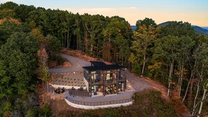 A modern two-story cabin with a deck, surrounded by dense forest. - Elevated Escapes by Carolina Mornings | Luxury w/PERFECT mountain views near AVL (Candler)