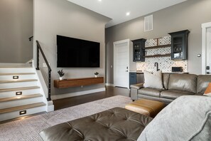 A modern living room with a flat-screen TV, a leather sofa, a coffee table, and a staircase with built-in lighting. - Elevated Escapes by Carolina Mornings | Luxury w/PERFECT mountain views near AVL (Candler)