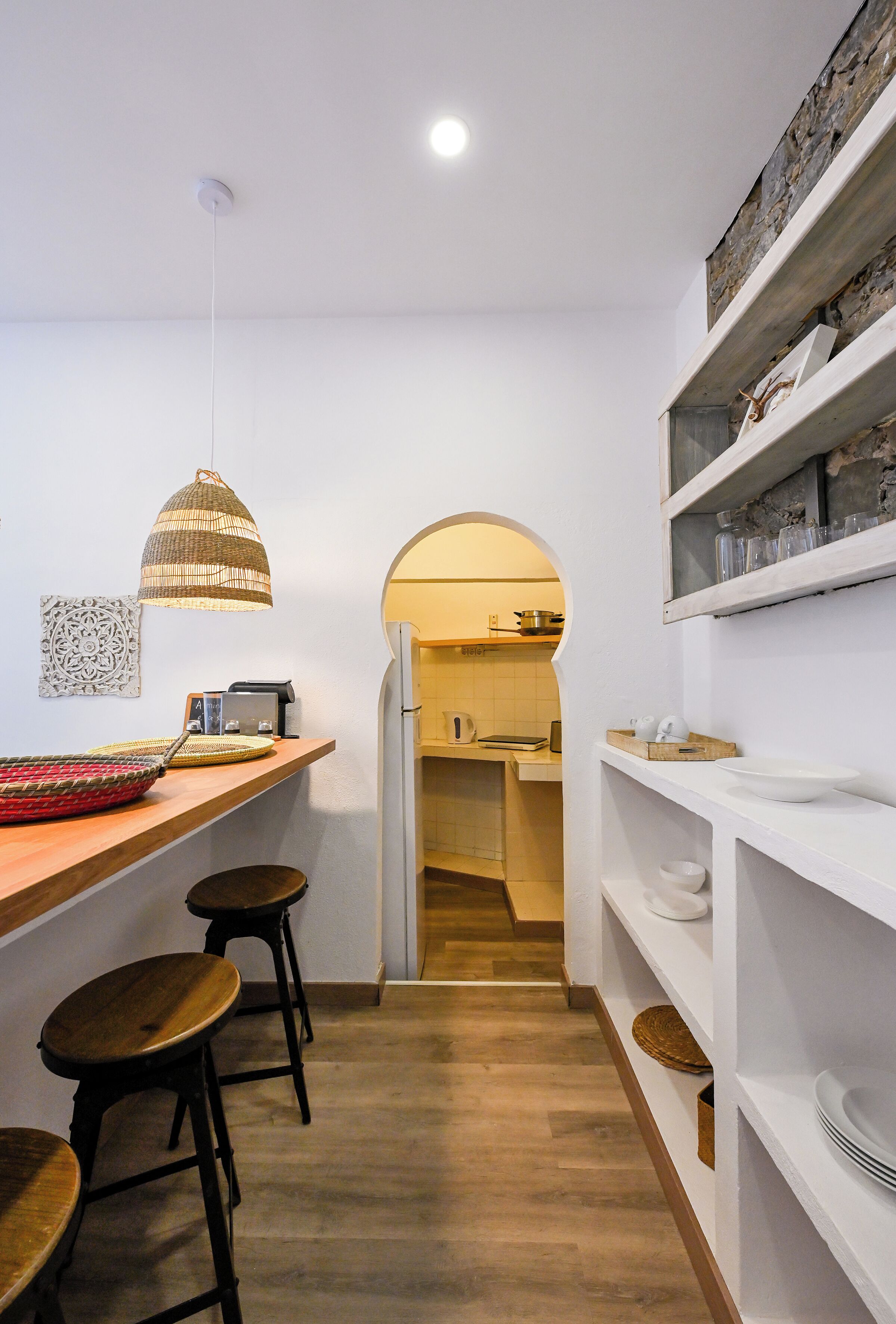 Une cuisine étroite avec un plan de travail en bois, des tabourets, un luminaire suspendu et une porte-fenêtre donnant sur un coin repas.