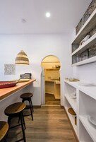 A narrow kitchen with a wooden countertop, stools, a hanging light fixture, and a doorway leading to a dining area.