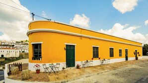 A yellow building with a green door, windows, and a paved area with chairs and potted plants. - Casa Amarela (Mértola)