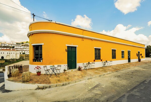A yellow building with a green door, windows, and a paved area with chairs and potted plants. - Casa Amarela (Mértola)