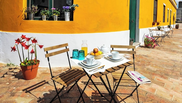 A yellow building with a green door, a window with potted plants, and a table set with plates, cups, and a menu. - Casa Amarela (Mértola)