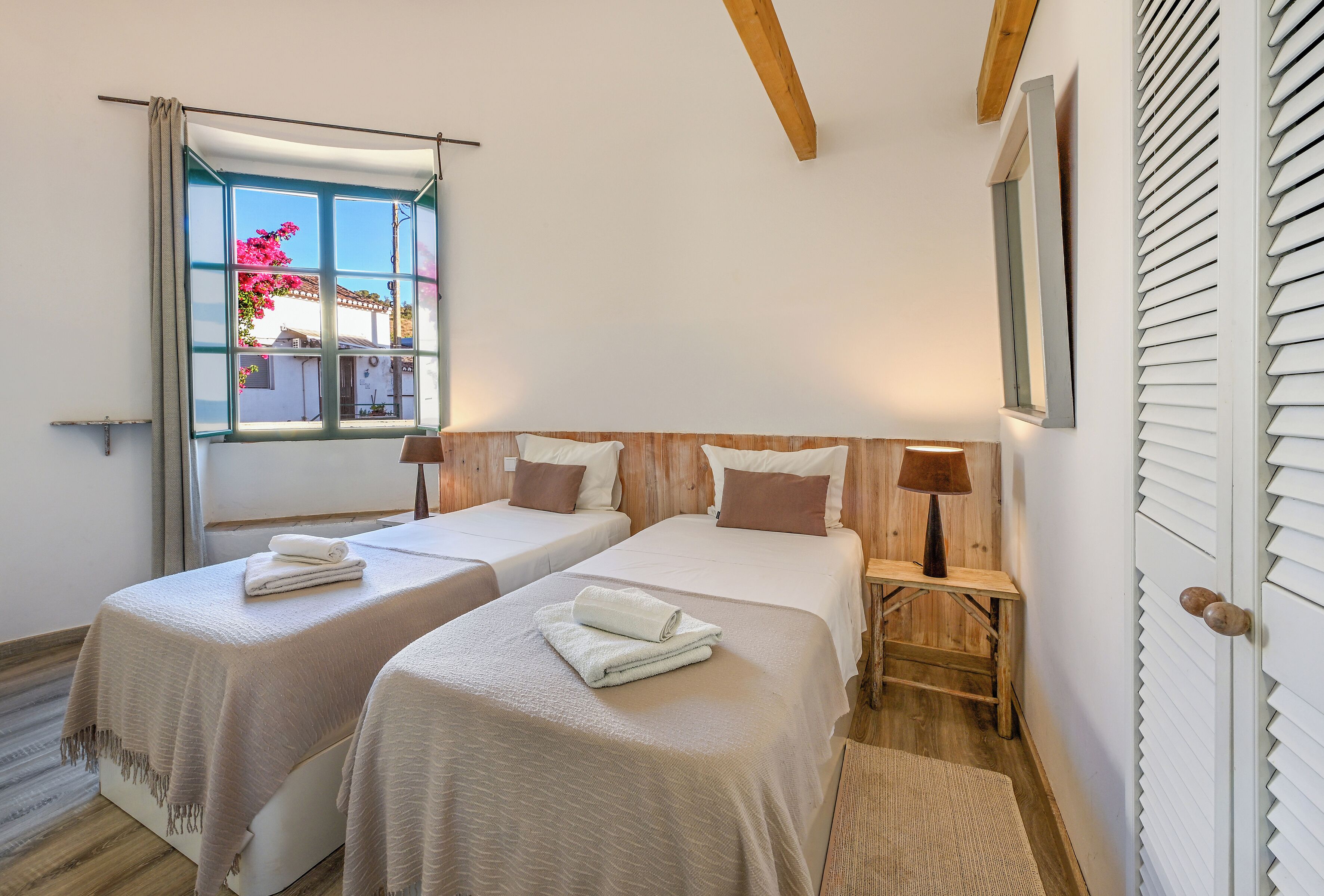 Quarto com duas camas, janela com cortinas, cabeceira de madeira, mesa de cabeceira com abajur e vista para um prédio.