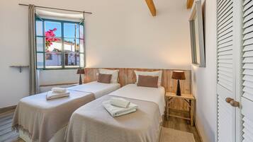 Quarto com duas camas, janela com cortinas, cabeceira de madeira, mesa de cabeceira com abajur e vista para um prédio.