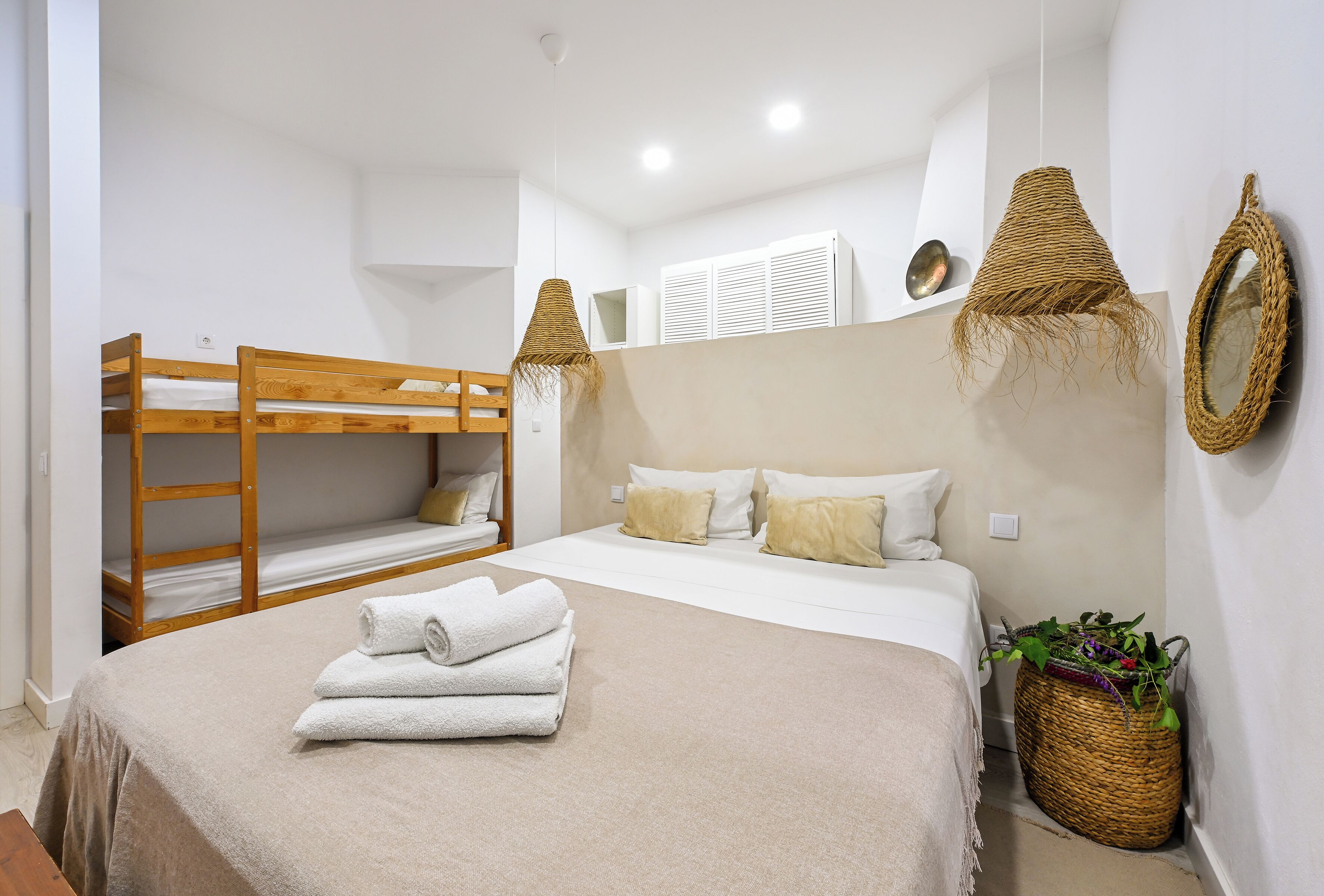 Un dormitorio con una cama doble, una cama individual con almohadas, una toalla enrollada, una cesta tejida con plantas y un espejo montado en la pared.