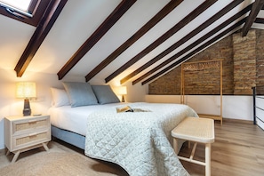 Een slaapkamer met een schuine plafond, een bed met quilt, nachtlampjes, een nachtkastje, een rieten stoel en een bakstenen muur.