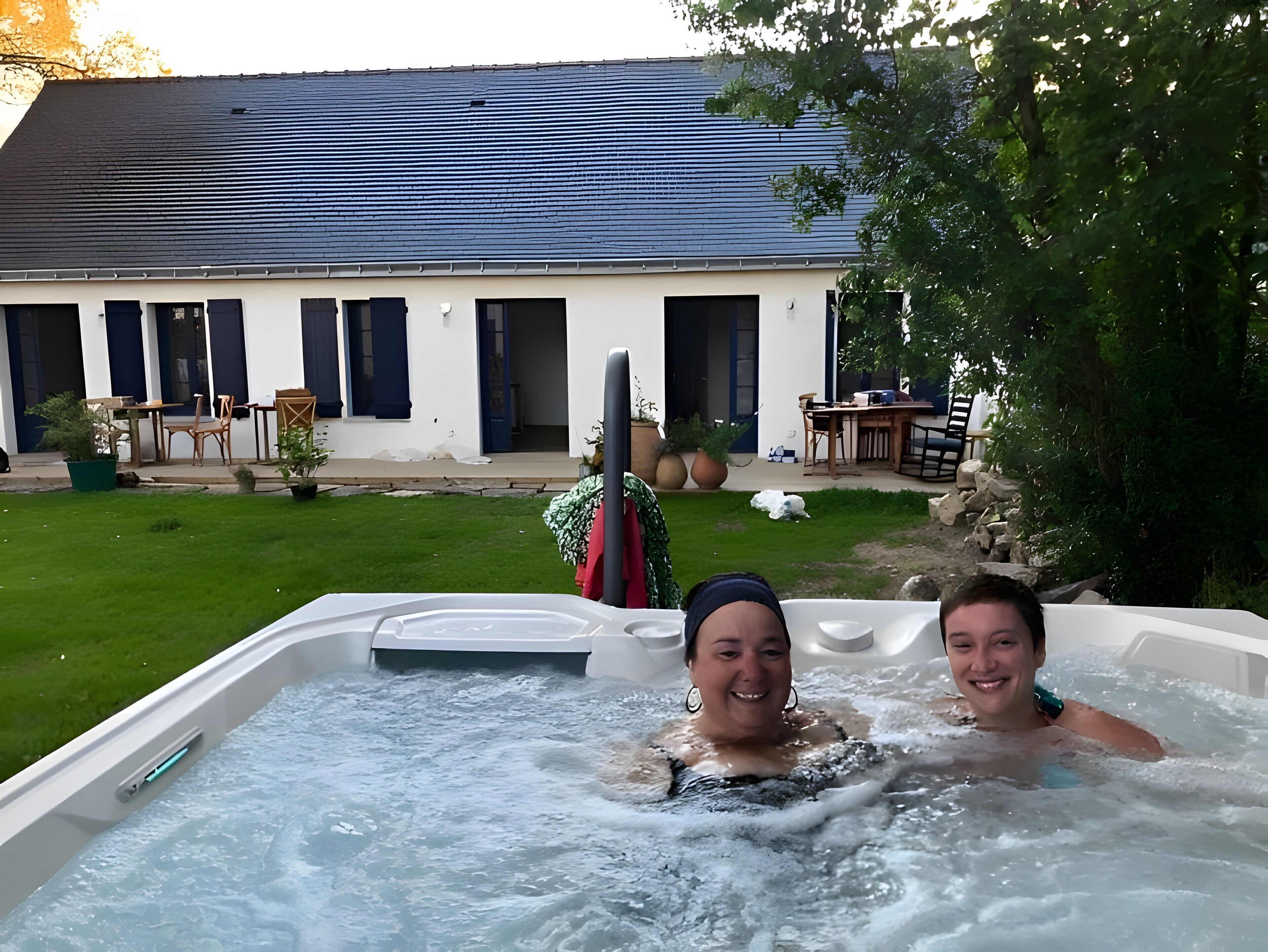 Un jacuzzi con dos personas, rodeado por un césped y un edificio con techo inclinado.