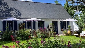 A two-story building with a grey tiled roof, a garden with various plants and flowers, and three umbrellas. - Bed and Breakfast 'Chambre 1 Coté Plage' with Private Terrace, Shared Garden and Wi-Fi (Arzon)