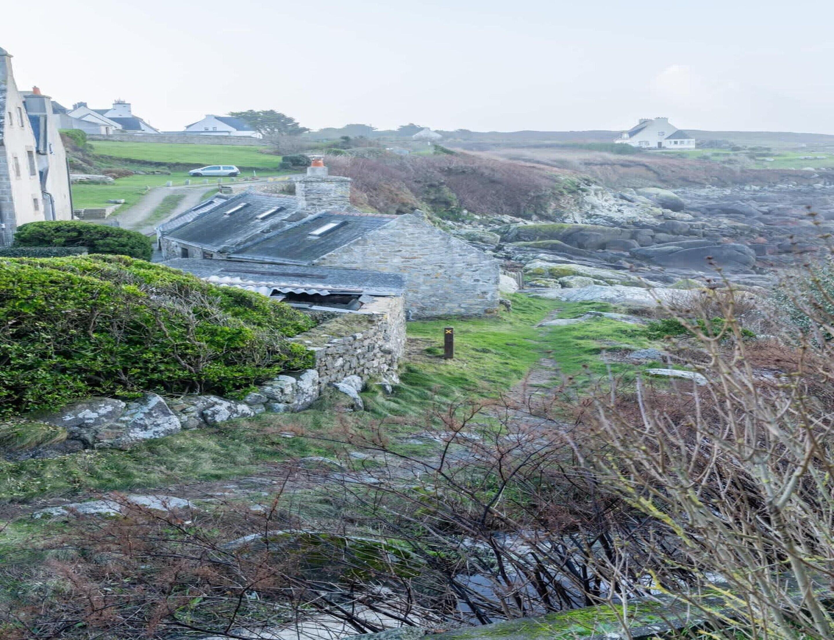 Ein Küstenhaus aus Stein mit geneigtem Dach, umgeben von felsigem Gelände und vereinzelter Vegetation.