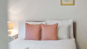 Un letto rifatto con cuscini bianchi e color pesca, una coperta a quadri e una lampada da comodino.