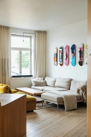 Ein modernes Wohnzimmer mit einer Couch, einem Hocker und einem Couchtisch. Ein Fenster mit Vorhängen und eine Wand mit Skateboard-Kunstwerken.