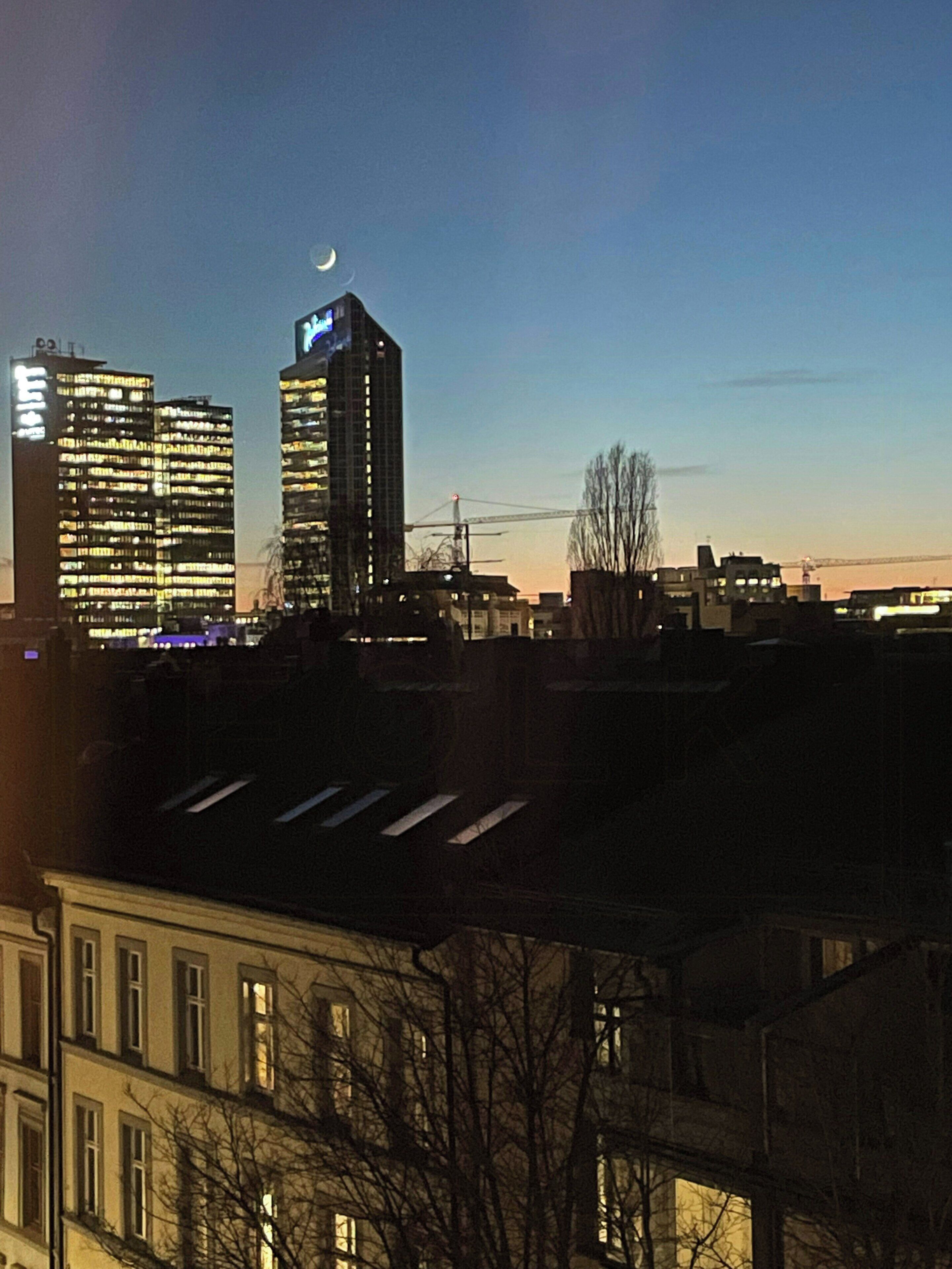 A city skyline at dusk with illuminated buildings, a crescent moon, and a bridge in the distance.
