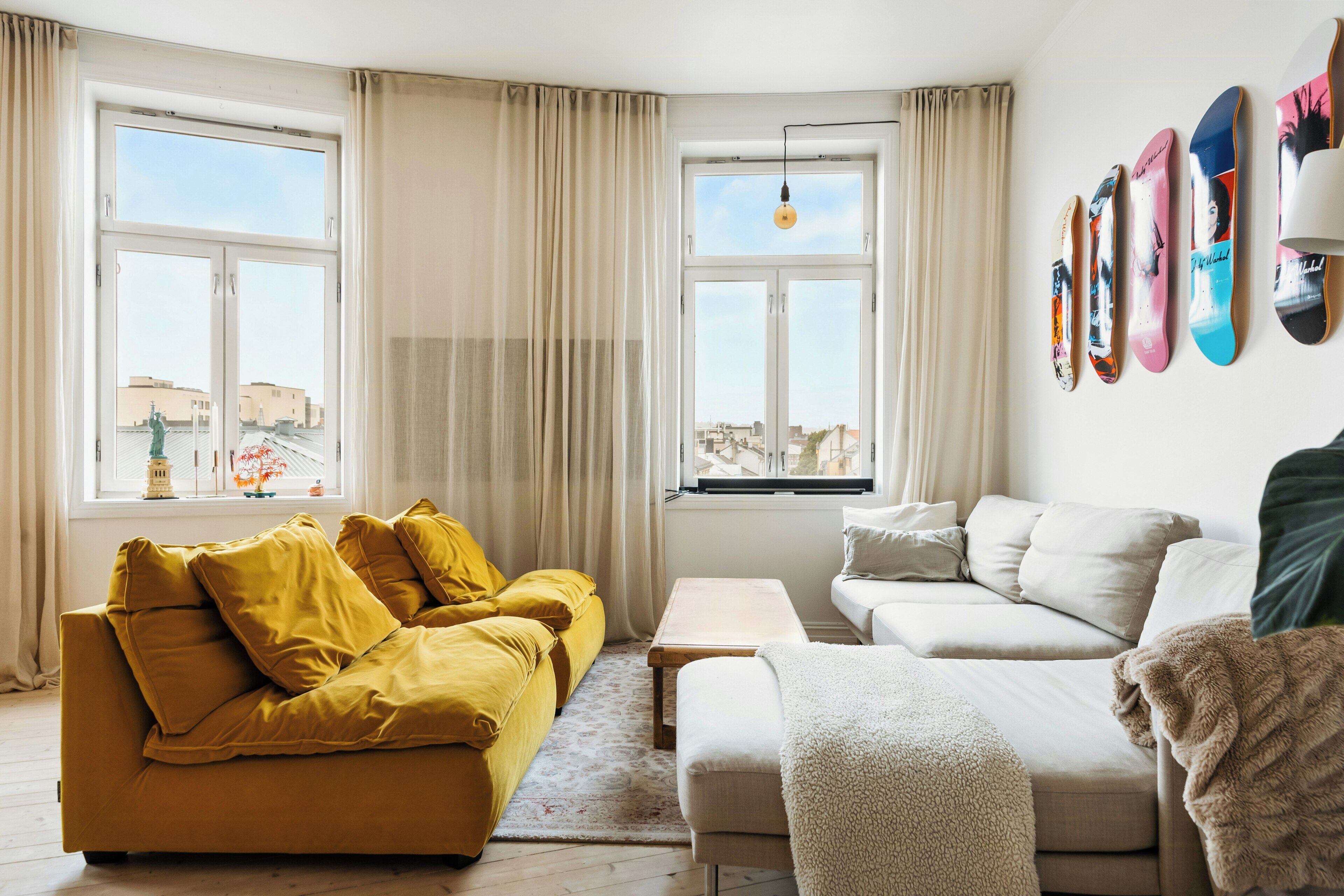 Una sala de estar con un sofá amarillo, un sofá blanco y una mesa de centro. Hay dos ventanas con cortinas y una pared decorada con cuadros.