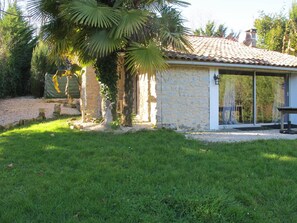 A stone building with a tiled roof, a covered patio, and a well-maintained lawn. - GITE DANS UN  ECRIN DE VERDURE à 25 MN DE L'OCEAN (Sainte-Marie-de-Gosse)