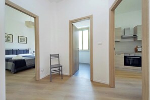 Ein Zimmer mit einem Bett, einem Sessel, einer Küchenzeile mit Schränken und einem Backofen sowie einer Tür zu einem weiteren Raum.