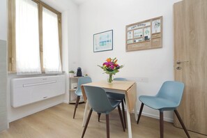 Una stanza con una porta in legno, una finestra con tende leggere, un tavolo con sedie, un termosifone, un quadro appeso al muro e una bacheca con diversi oggetti.