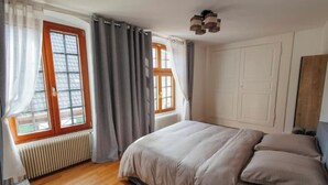 Un dormitorio con cama, dos ventanas con cortinas, un radiador y suelo de madera.