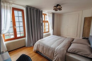 A bedroom with a bed, two windows with curtains, a radiator, and a wooden floor. - La Cure Apartment (Wintzenheim)