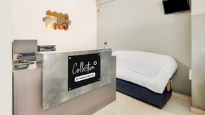 Un mostrador de recepción de hotel con un letrero que dice 'Collection', un ventilador de techo, un televisor montado y una cama con una colcha blanca.